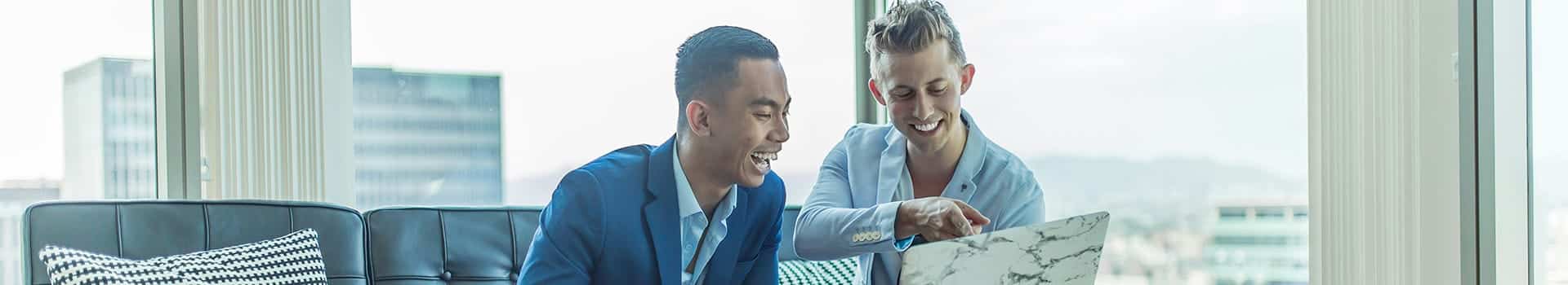 Två personer i kostym sitter på en soffa och tittar på en bärbar dator, båda ler.